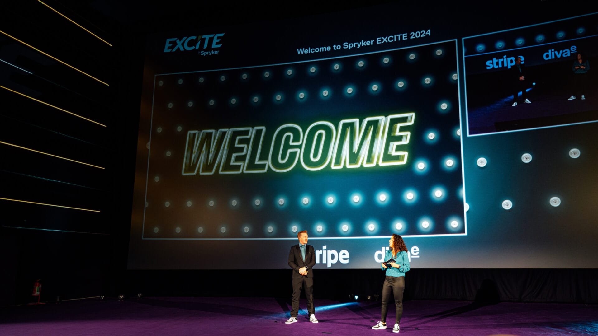 Two people stand on a stage in front of a large screen displaying "WELCOME" at the Spryker EXCITE 2024 event, setting the tone for what’s to come at Spryker EXCITE 2025.