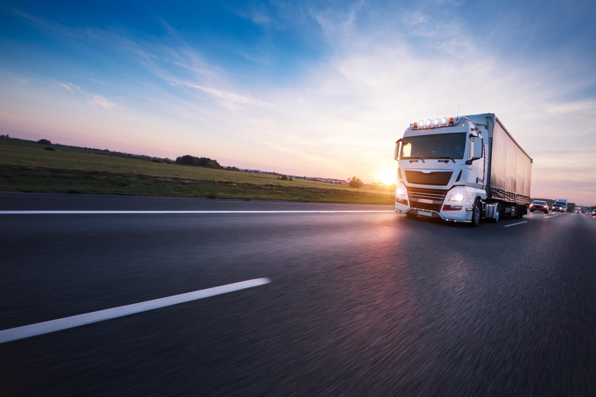 A large white truck with a trailer is driving on a highway at sunset. The sky is a gradient of blue and orange, and the road stretches into the distance with cars in the far lanes. Open fields and distant trees line the sides of the highway.