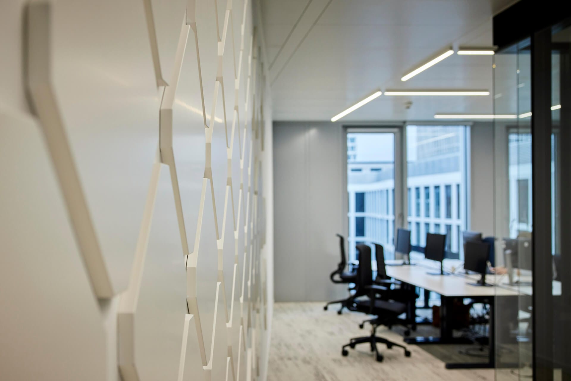 Modern office with several empty desks and chairs, a glass wall, computers, and natural light streaming through large windows; foreground features a geometric wall design—an inspiring workspace at Daimler Truck.